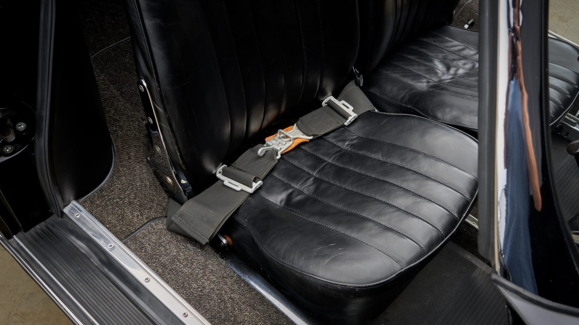 A vintage two-point lap belt with a metal lift-latch buckle lying across a black leather bucket seat.
