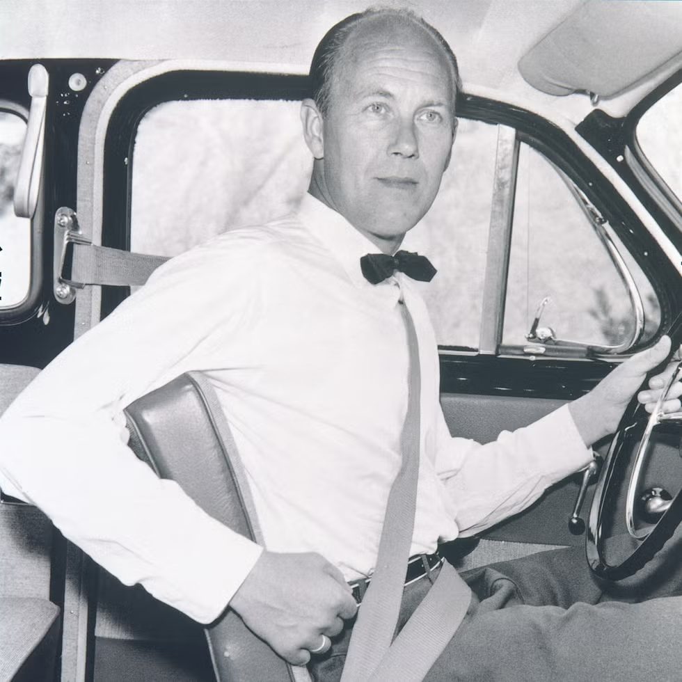 Black-and-white photo of Nils Bohlin, wearing a bow tie, sitting in a car with one hand on the wheel and the diagonal seat belt strap across his chest.