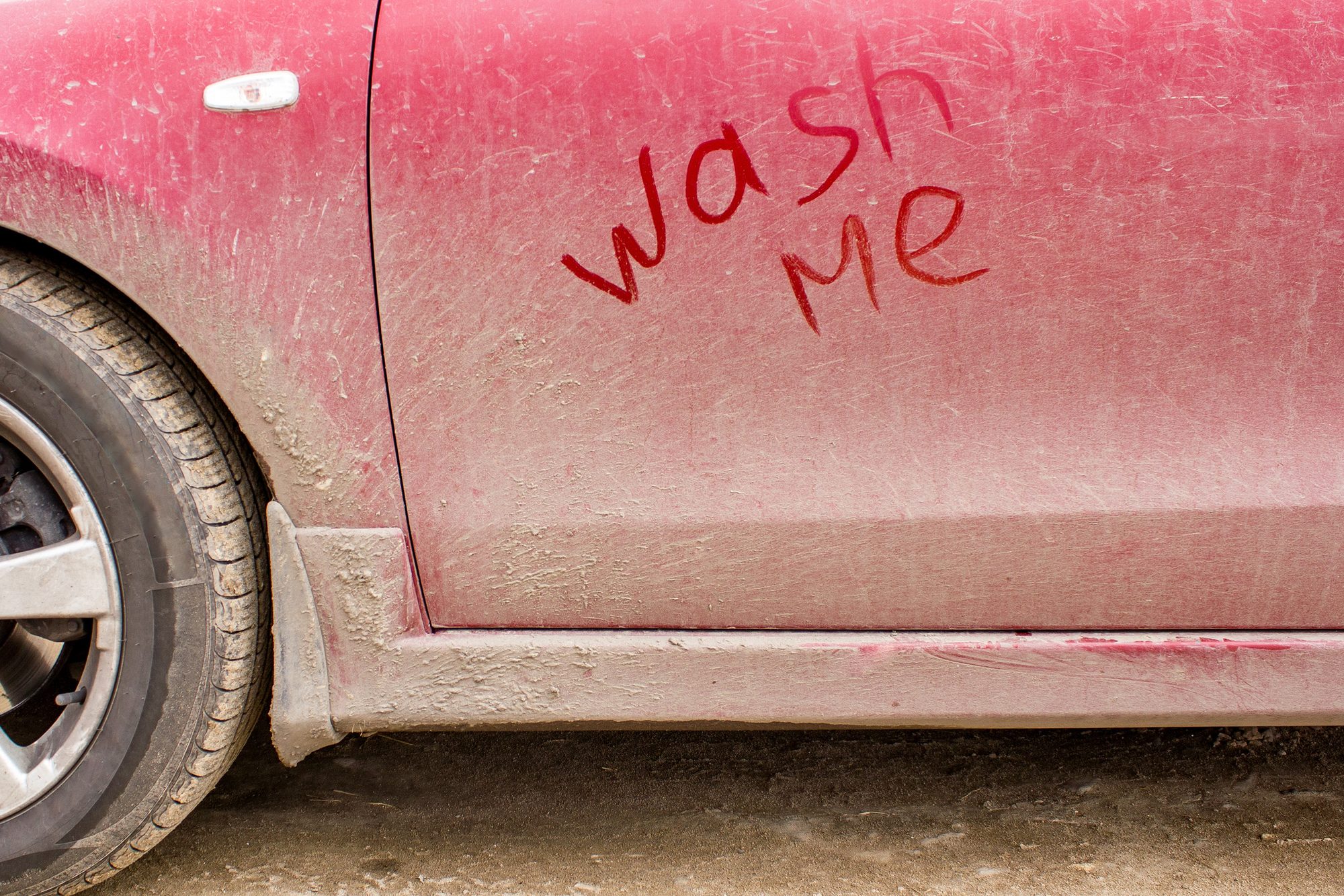 A dust-caked red car door with the words wash me written in the grime, mud caked along the rocker panel and wheel arch
