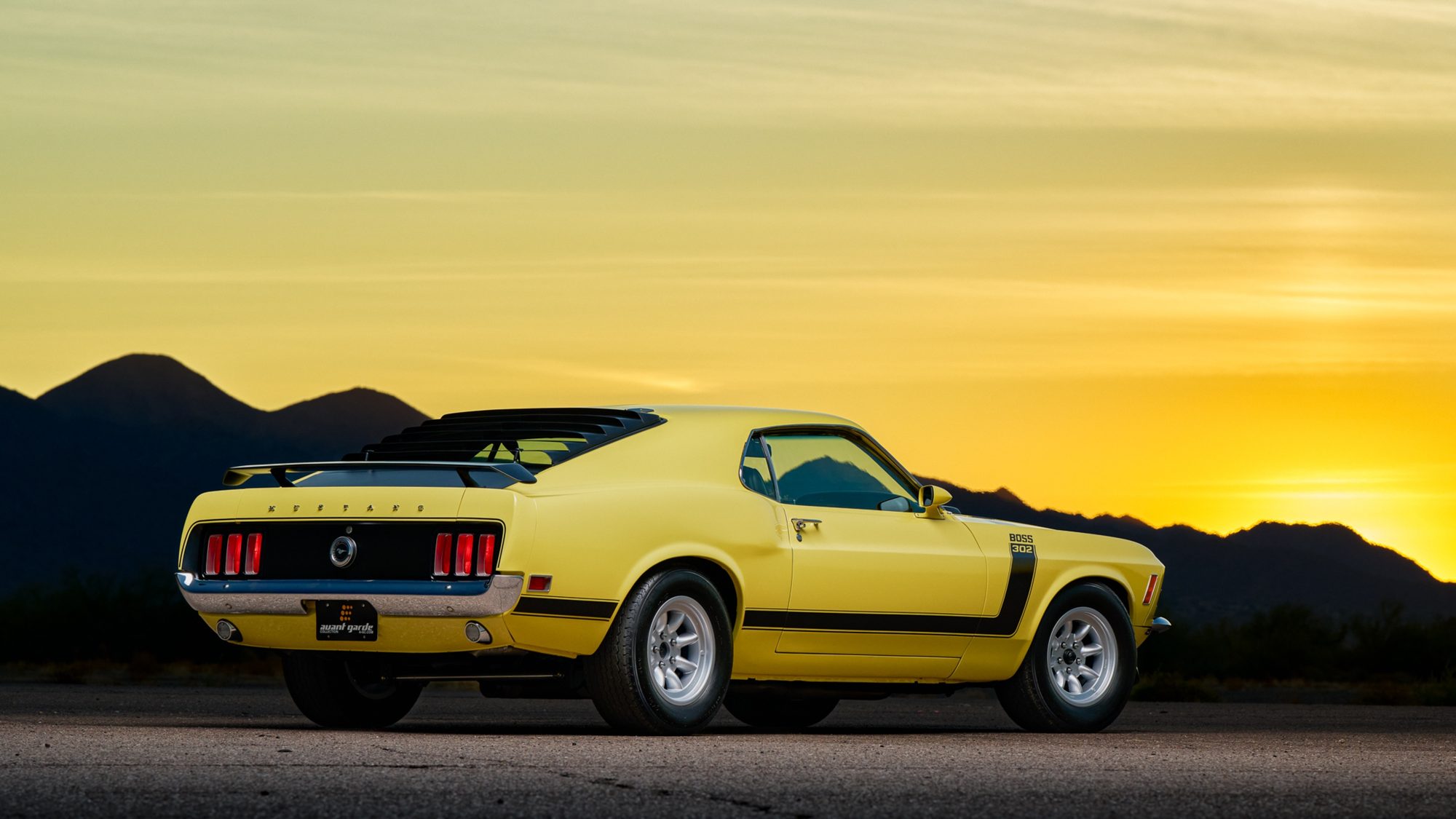 A yellow 1970 Ford Mustang Boss 302 with black side stripes, rear window louvers and a rear wing, seen from the rear three-quarter angle at sunset with mountains in the background