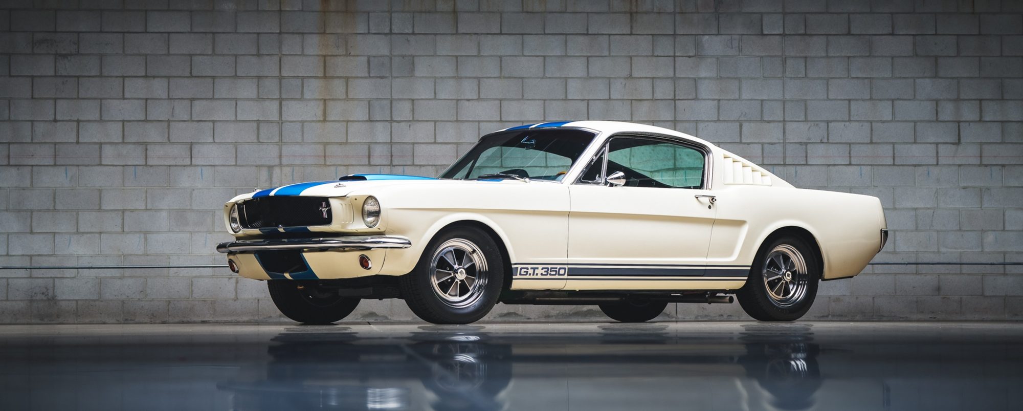 A white 1965 Shelby GT350 fastback with blue racing stripes and G.T. 350 rocker stripes, parked against a grey concrete block wall on a reflective floor