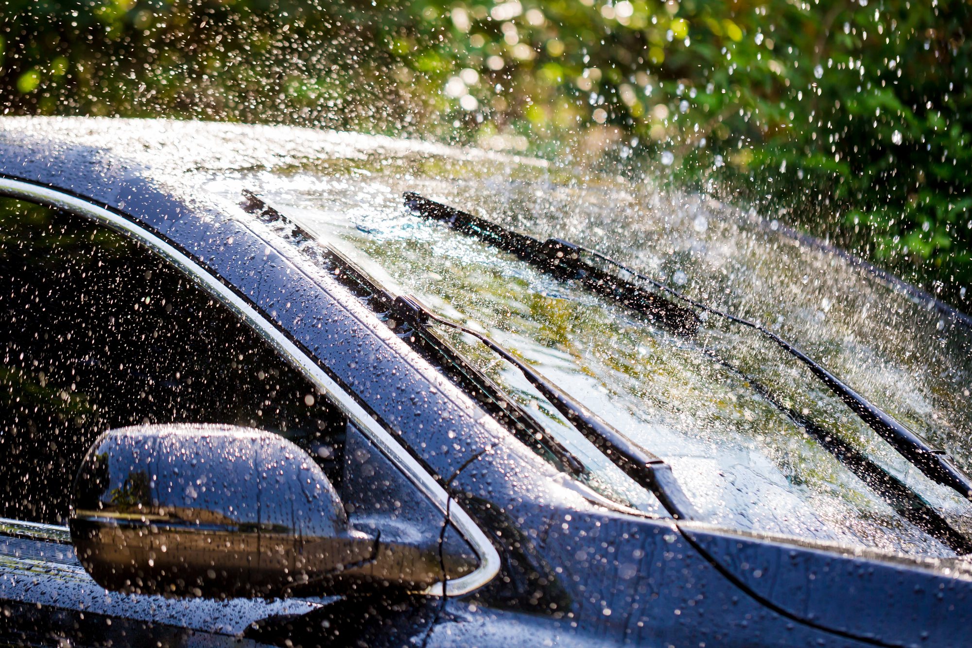 Water droplets spray across the windshield and side mirror of a dark car, its two wiper blades lying on the wet glass, with sunlit green trees behind.