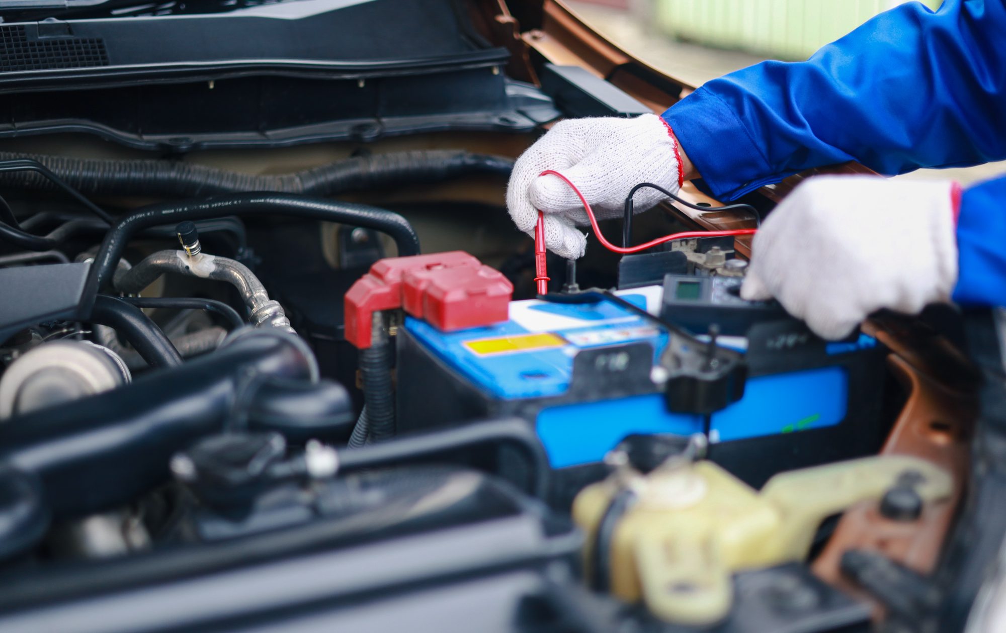 A mechanic in white work gloves touches a multimeter's red probe to a car battery terminal under the hood