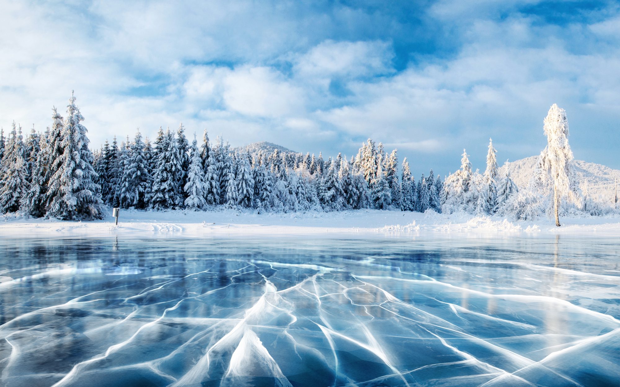 Cracked blue ice on a frozen lake, with snow-laden pines and hills beyond under a cloudy blue sky