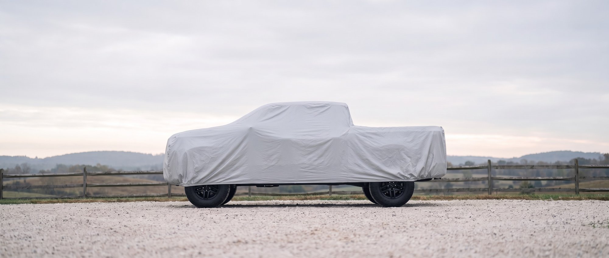 A full-size pickup under a fitted silver outdoor cover on a gravel ranch drive, fence and hills behind