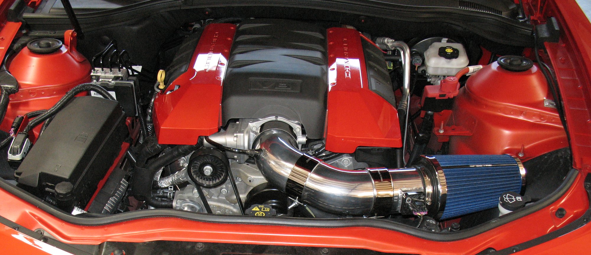 An aftermarket intake in the engine bay of a red Chevrolet Camaro SS: polished aluminum tubes joined by silicone couplers run from a blue cone air filter to the throttle body