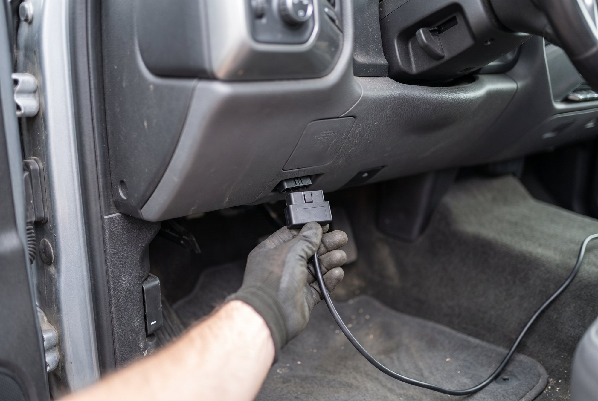 A gloved hand plugging a black cable into the OBD-II diagnostic port under the dashboard of a pickup