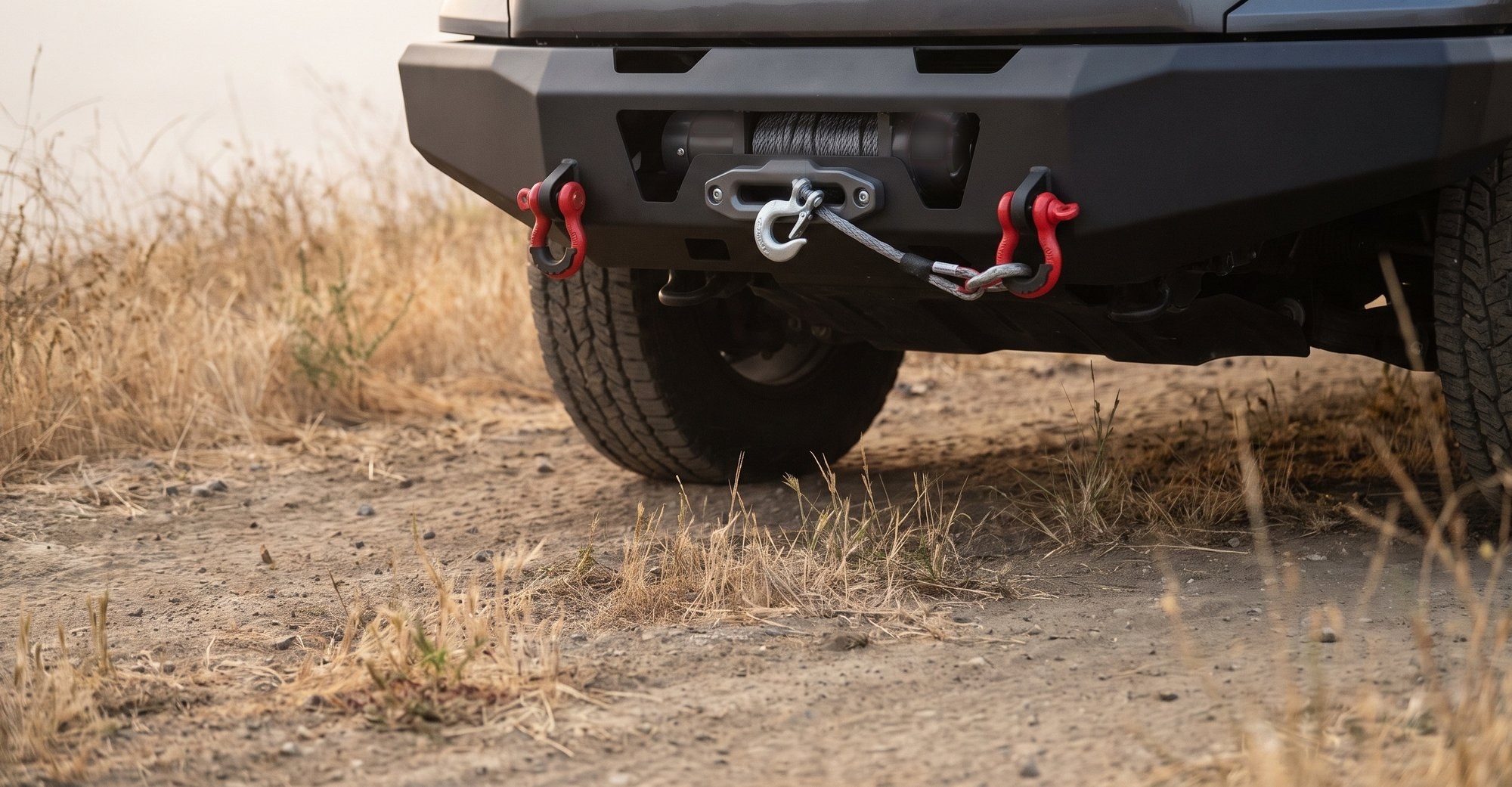 The front of a grey pickup fitted with a black steel recovery bumper, two red D-ring shackles and a center winch with synthetic rope and a hook, on a dirt trail