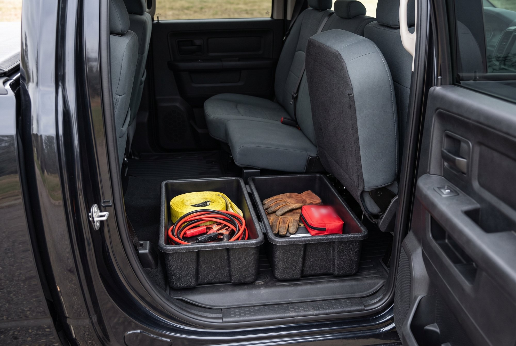 The rear cab of a crew-cab pickup with the bench cushion folded up, showing two black storage bins on the floor that hold a tow strap, jumper cables, work gloves and a first-aid pouch