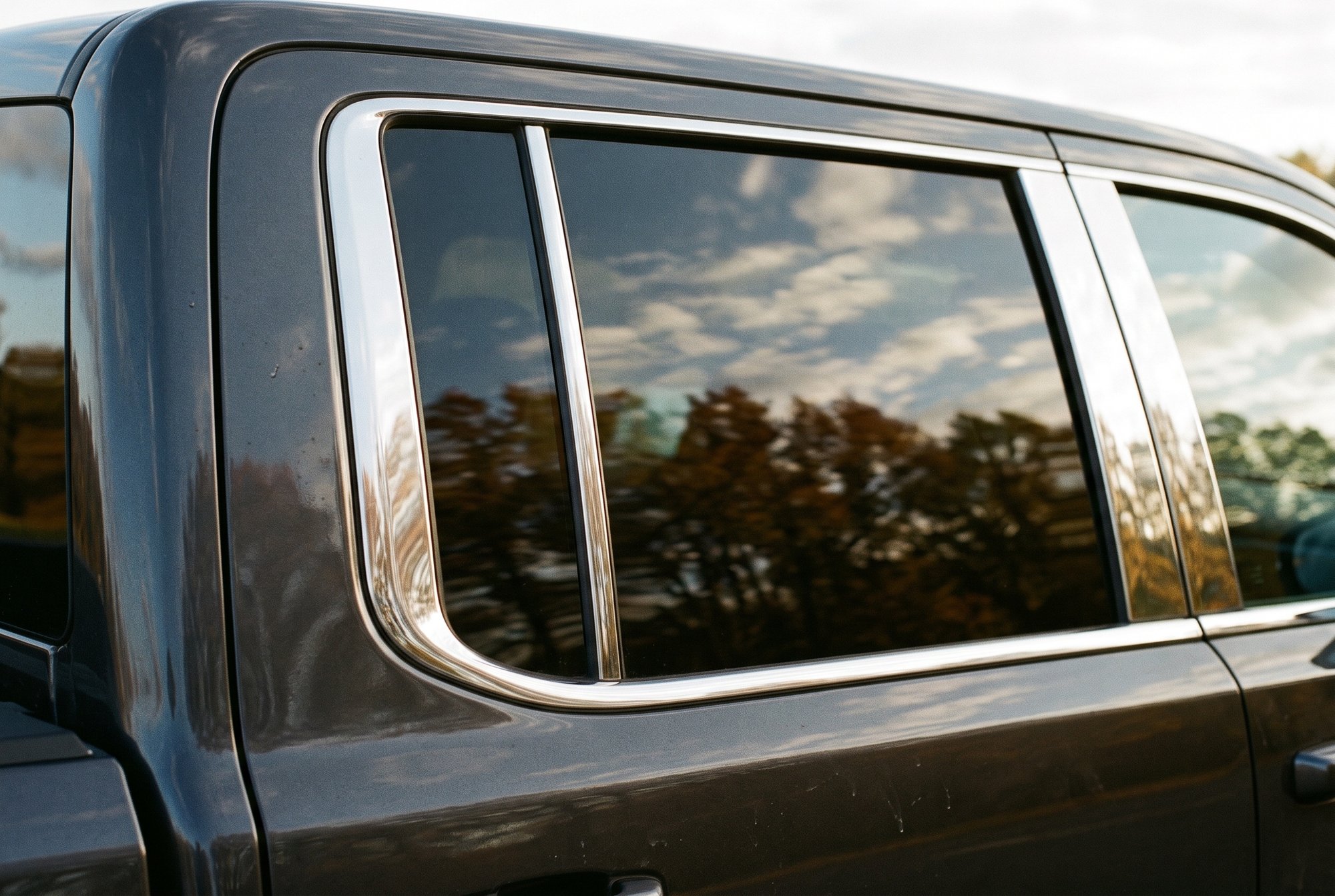 The rear door window of a charcoal grey pickup, framed by polished stainless steel trim that reflects the sky