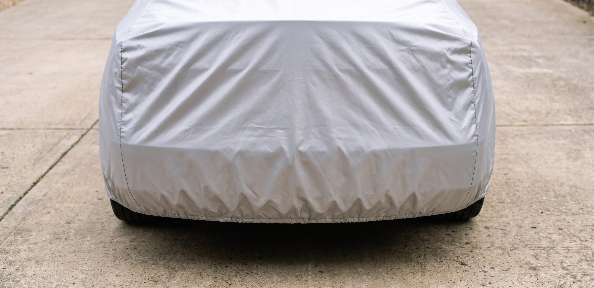 The rear of a sedan under a silver cover whose elastic hem is drawn in under the bumper, lying close to the body on a concrete driveway