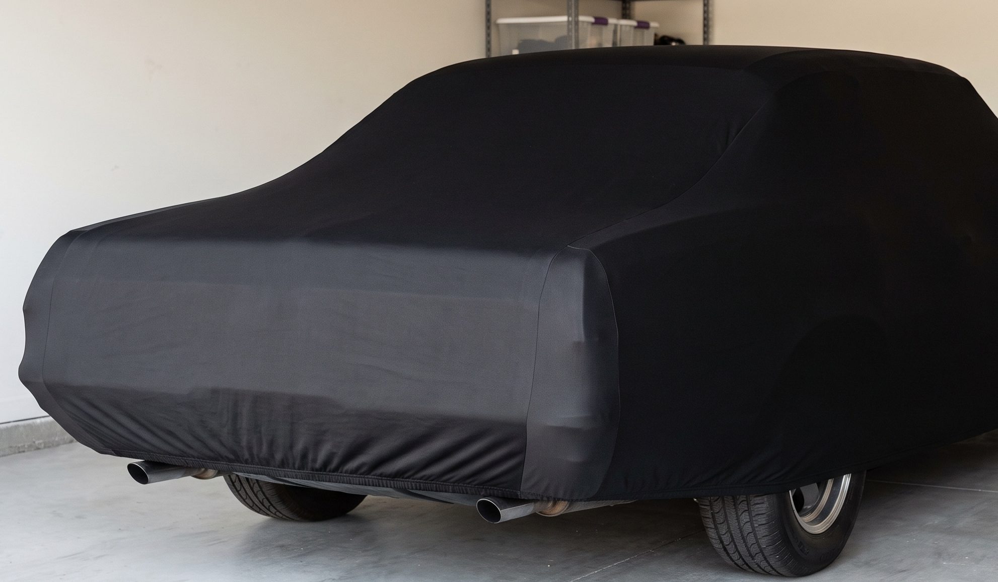 The rear of a classic coupe in a tidy garage wearing a close-fitting black stretch indoor cover, hem drawn in under the bumper, exhaust tips below the cover
