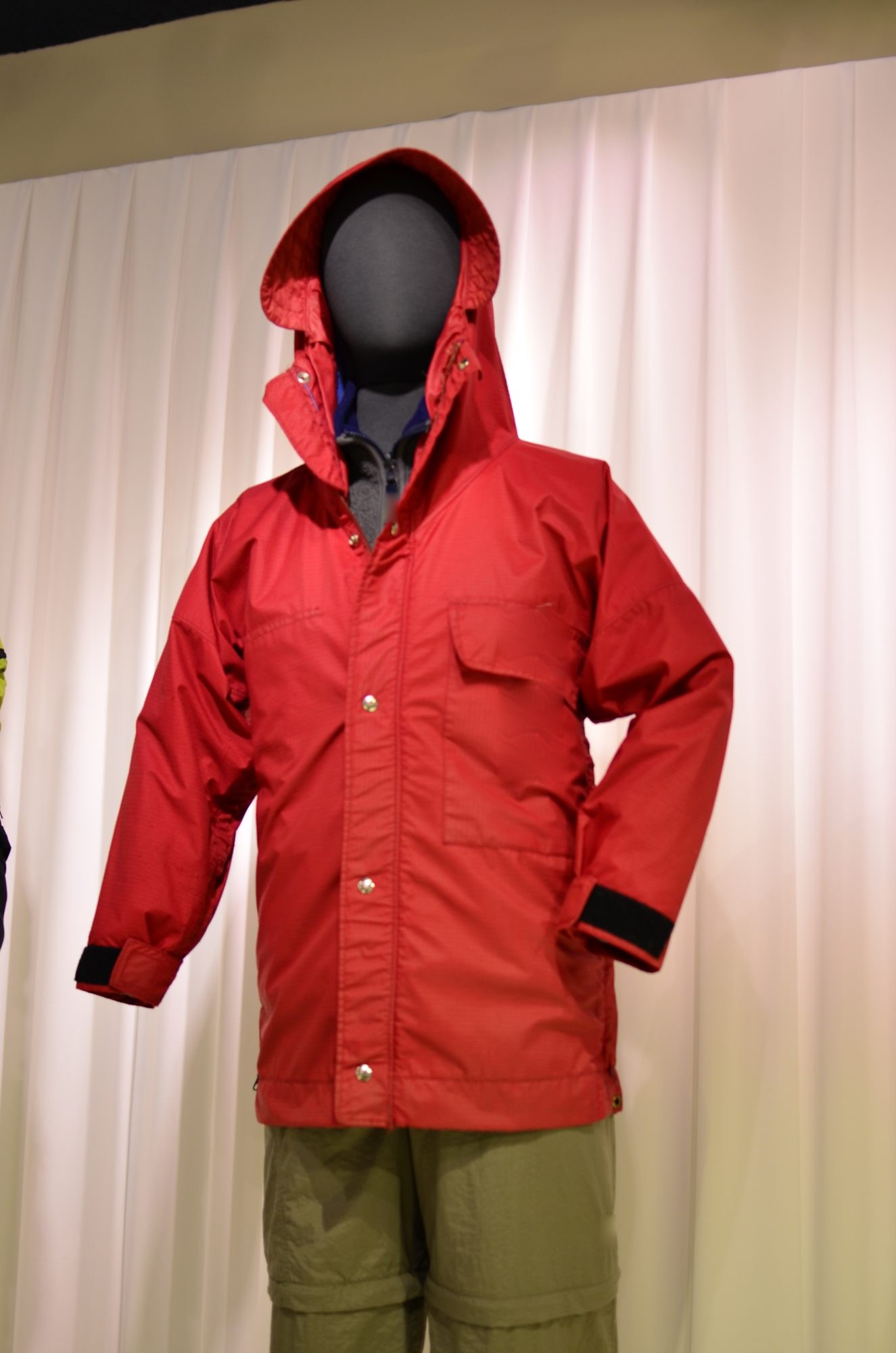 A red hooded rain jacket with a flap chest pocket on a mannequin against a white curtain