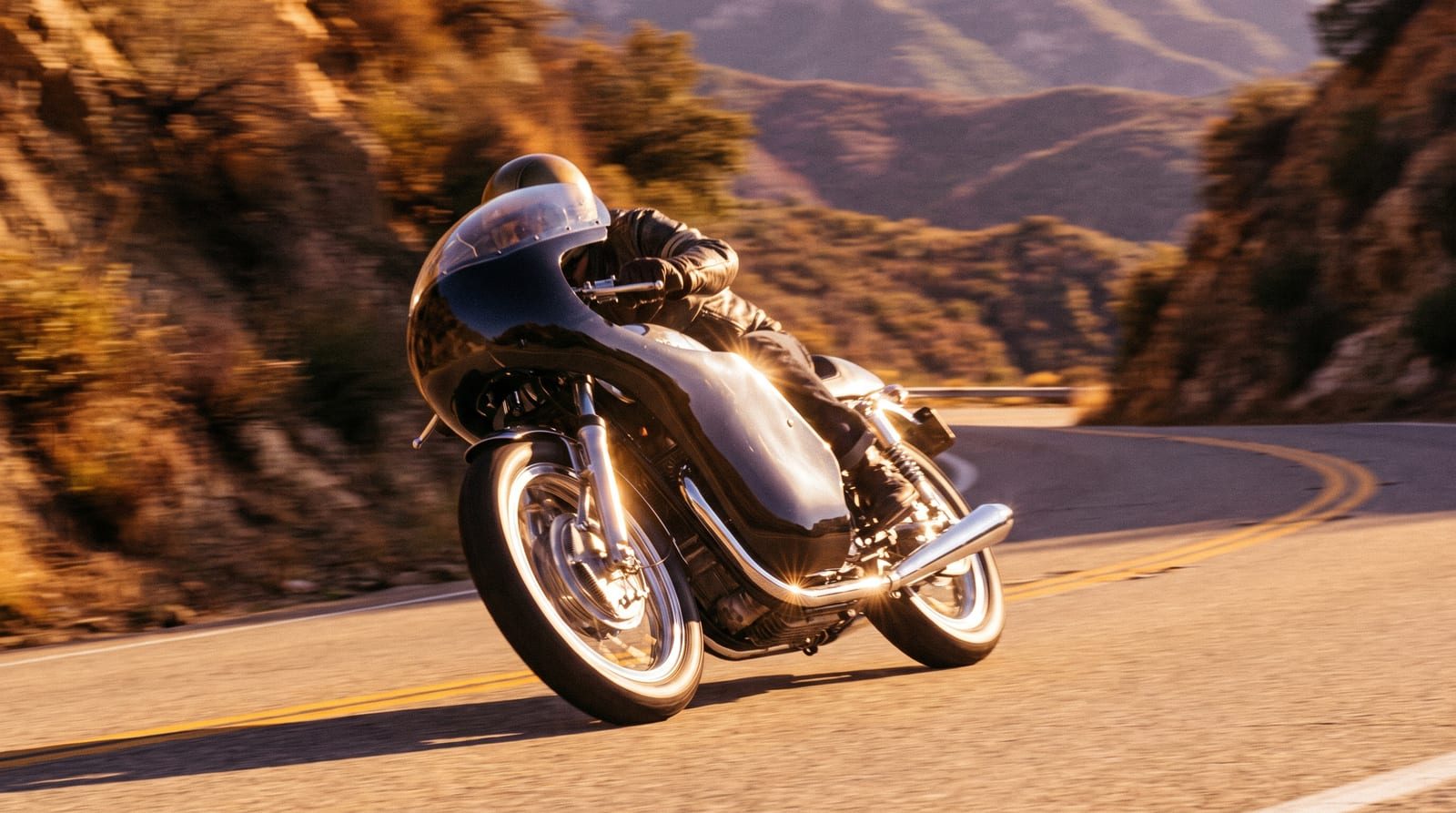 A rider leaning a café racer through a canyon bend in golden light.